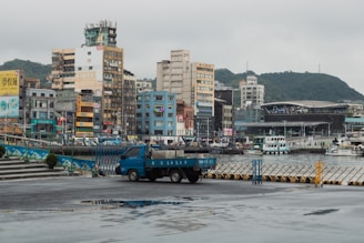 An urban waterfront scene featuring a variety of mid-rise buildings along the shoreline. A blue delivery truck is parked in the foreground on a wet asphalt surface that reflects some of the surroundings. The area has visible signage in multiple languages, and there is a harbor with several boats docked. Green hills and a cloudy sky form the backdrop, adding a sense of depth to the setting.