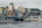 An urban waterfront scene featuring a variety of mid-rise buildings along the shoreline. A blue delivery truck is parked in the foreground on a wet asphalt surface that reflects some of the surroundings. The area has visible signage in multiple languages, and there is a harbor with several boats docked. Green hills and a cloudy sky form the backdrop, adding a sense of depth to the setting.