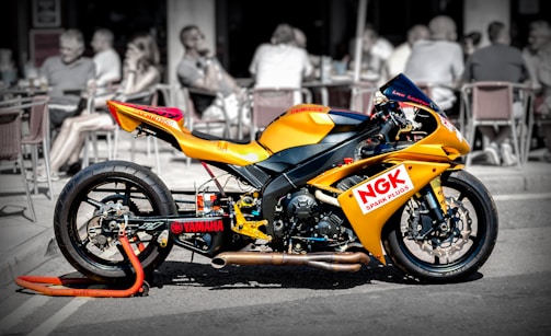 A bright yellow Yamaha racing motorcycle is parked on a street with a cafe and people blurred in the background. The bike is positioned on an orange stand, displaying logos like NGK Spark Plugs prominently. The focus is on the motorcycle, contrasting with the black and white effect on the backdrop.