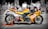 A bright yellow Yamaha racing motorcycle is parked on a street with a cafe and people blurred in the background. The bike is positioned on an orange stand, displaying logos like NGK Spark Plugs prominently. The focus is on the motorcycle, contrasting with the black and white effect on the backdrop.