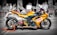 A bright yellow Yamaha racing motorcycle is parked on a street with a cafe and people blurred in the background. The bike is positioned on an orange stand, displaying logos like NGK Spark Plugs prominently. The focus is on the motorcycle, contrasting with the black and white effect on the backdrop.