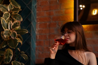 A woman with glasses enjoys a drink from a wine glass in a warmly lit room. The background features a brick wall and green plants.
