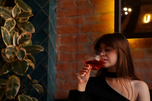 A woman with glasses enjoys a drink from a wine glass in a warmly lit room. The background features a brick wall and green plants.