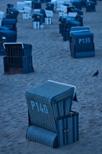 Several beach chairs with numbers painted on them are arranged in rows on the sandy beach. The chairs predominantly feature blue and white striped designs. A small bird is seen on the sand in the foreground.