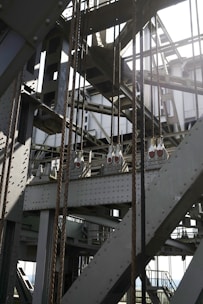 An intricate structure of metal beams and cables featuring industrial elements such as riveted steel girders and wires in a geometric pattern. Light and shadows create a dramatic effect across the metallic surfaces.