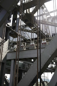 An intricate structure of metal beams and cables featuring industrial elements such as riveted steel girders and wires in a geometric pattern. Light and shadows create a dramatic effect across the metallic surfaces.