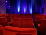 A dimly lit theater features rows of red plush seats. Soft blue lighting bathes the curtain on the stage, adding a serene ambiance. The theater is empty, contributing to a quiet and intimate atmosphere.