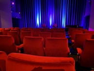 A dimly lit theater features rows of red plush seats. Soft blue lighting bathes the curtain on the stage, adding a serene ambiance. The theater is empty, contributing to a quiet and intimate atmosphere.