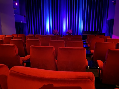 A dimly lit theater features rows of red plush seats. Soft blue lighting bathes the curtain on the stage, adding a serene ambiance. The theater is empty, contributing to a quiet and intimate atmosphere.