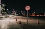 A dimly lit urban walkway at night features modern streetlights illuminating the path. In the foreground, a traffic sign indicates no cycling, mounted on a pole. Traffic cones are positioned on the dirt adjacent to the path. Low-rise buildings and trees with ambient lighting create an urban backdrop.