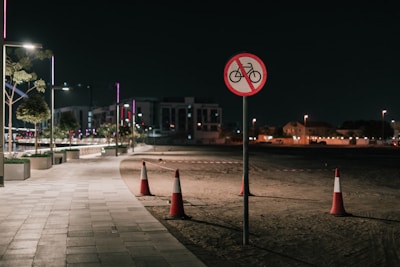 A dimly lit urban walkway at night features modern streetlights illuminating the path. In the foreground, a traffic sign indicates no cycling, mounted on a pole. Traffic cones are positioned on the dirt adjacent to the path. Low-rise buildings and trees with ambient lighting create an urban backdrop.