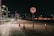 A dimly lit urban walkway at night features modern streetlights illuminating the path. In the foreground, a traffic sign indicates no cycling, mounted on a pole. Traffic cones are positioned on the dirt adjacent to the path. Low-rise buildings and trees with ambient lighting create an urban backdrop.