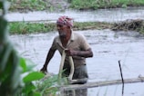 A person is standing in a flooded or swampy area, wearing a headscarf and a worn shirt. They are holding a long piece of cloth or rope and appear to be working in the water. The surroundings consist of wet mud and green vegetation.