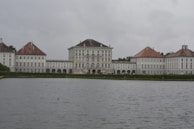 A large historical palace with a symmetrical architectural design, featuring a central building flanked by two wings. The structure is positioned near a body of water with a cloudy sky in the background. The palace has a classic European style, with multiple windows and red-tiled roofs.