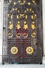An ornate, wrought-iron gate featuring intricate designs with floral and spiral patterns. The gate is accented with gold-colored leaves and decorative elements, set against a red background.