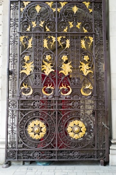 An ornate, wrought-iron gate featuring intricate designs with floral and spiral patterns. The gate is accented with gold-colored leaves and decorative elements, set against a red background.