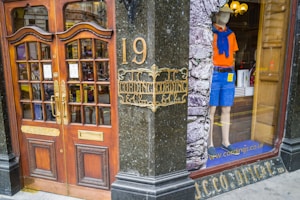 A storefront with ornate wooden doors featuring glass panels and brass handles, situated at number 19. Next to the entrance, a mannequin is displayed in the window wearing casual summer attire: an orange shirt, blue shorts, a blue sweater tied around the shoulders, and slip-on shoes. The window has an interior display with a rustic brick texture.