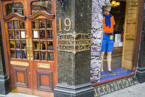 A storefront with ornate wooden doors featuring glass panels and brass handles, situated at number 19. Next to the entrance, a mannequin is displayed in the window wearing casual summer attire: an orange shirt, blue shorts, a blue sweater tied around the shoulders, and slip-on shoes. The window has an interior display with a rustic brick texture.