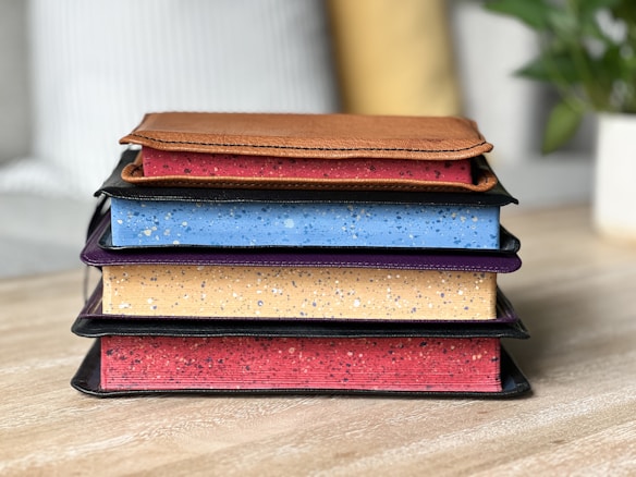 A stack of four books lies on a wooden surface. Each book has a unique speckled pattern on its edge, with colors ranging from red, blue, beige, and purple. On top of the stack is a brown leather-bound diary or notebook. In the background, there is a blurred view of a sofa with striped cushions and a green plant in a pot.