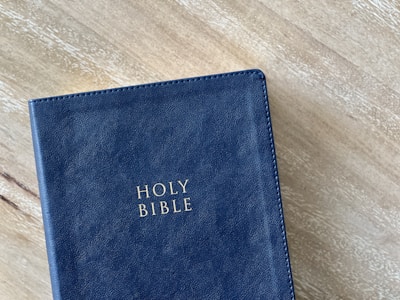 A leather-bound book titled 'Holy Bible' with gold lettering is placed on a wooden surface. The cover is dark blue and has a textured finish, giving it an elegant appearance.