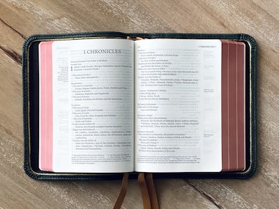 An open book with the title '1 Chronicles' at the top of the left page, displaying several columns of text. The book is placed on a wooden surface, and a bookmark ribbon is visible.