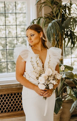 A woman wearing an elegant white bridal gown holds a bouquet of white flowers. She stands near a large window with a simple grid pattern, and lush green plants are visible in the background. Her hair is styled neatly, and she is wearing subtle makeup and earrings.