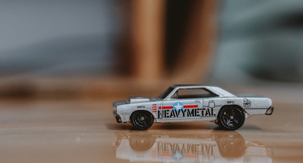 A small toy car with a retro design, featuring a metallic body with colorful decals including text and a star emblem. The surface is reflective, and the background is blurred, suggesting a shallow depth of field.