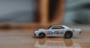 A small toy car with a retro design, featuring a metallic body with colorful decals including text and a star emblem. The surface is reflective, and the background is blurred, suggesting a shallow depth of field.