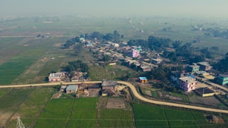 A small rural village nestled among expansive green fields. The area is dotted with clusters of colorful buildings and winding dirt roads, surrounded by lush vegetation and sparse trees. The hazy sky suggests a typical day in the countryside.