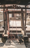 A rustic well with a wooden frame and pulley system. Red ropes are attached to a metal bucket, which rests on the stone slab. The background includes a courtyard with stone walls and stairs, enhancing the rural ambiance.