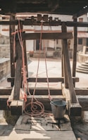A rustic well with a wooden frame and pulley system. Red ropes are attached to a metal bucket, which rests on the stone slab. The background includes a courtyard with stone walls and stairs, enhancing the rural ambiance.