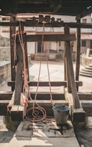 A rustic well with a wooden frame and pulley system. Red ropes are attached to a metal bucket, which rests on the stone slab. The background includes a courtyard with stone walls and stairs, enhancing the rural ambiance.