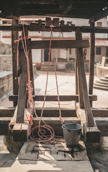 A rustic well with a wooden frame and pulley system. Red ropes are attached to a metal bucket, which rests on the stone slab. The background includes a courtyard with stone walls and stairs, enhancing the rural ambiance.