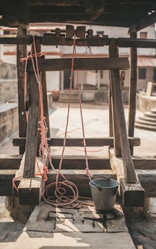 A rustic well with a wooden frame and pulley system. Red ropes are attached to a metal bucket, which rests on the stone slab. The background includes a courtyard with stone walls and stairs, enhancing the rural ambiance.