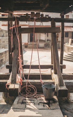 A rustic well with a wooden frame and pulley system. Red ropes are attached to a metal bucket, which rests on the stone slab. The background includes a courtyard with stone walls and stairs, enhancing the rural ambiance.