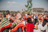 A large group of people participate in an outdoor festival or cultural event. Many individuals wear colorful, traditional attire with vibrant patterns, and some hold musical instruments. The atmosphere is lively and communal, with people gathered closely together, and a tree adorned with flags or decorations stands prominently in the center. In the background, a modern building is under construction against a backdrop of mountains and blue sky.