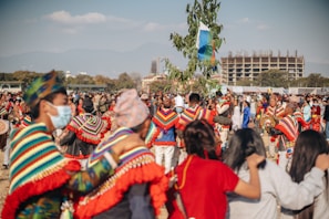 A large group of people participate in an outdoor festival or cultural event. Many individuals wear colorful, traditional attire with vibrant patterns, and some hold musical instruments. The atmosphere is lively and communal, with people gathered closely together, and a tree adorned with flags or decorations stands prominently in the center. In the background, a modern building is under construction against a backdrop of mountains and blue sky.