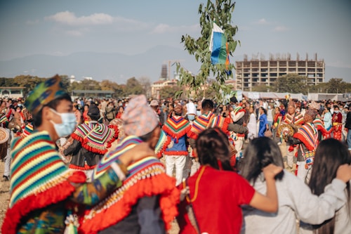 A large group of people participate in an outdoor festival or cultural event. Many individuals wear colorful, traditional attire with vibrant patterns, and some hold musical instruments. The atmosphere is lively and communal, with people gathered closely together, and a tree adorned with flags or decorations stands prominently in the center. In the background, a modern building is under construction against a backdrop of mountains and blue sky.