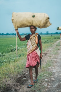 A person walking along a rural path carrying a large sack on their head. They are dressed in traditional clothing, with a multicolored shawl and a red skirt. The background features green fields under a clear blue sky, evoking a rural, agricultural setting.