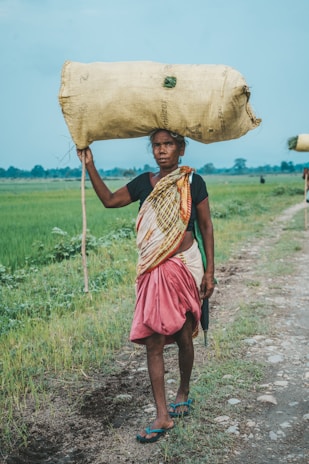 A person walking along a rural path carrying a large sack on their head. They are dressed in traditional clothing, with a multicolored shawl and a red skirt. The background features green fields under a clear blue sky, evoking a rural, agricultural setting.