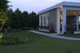 A modern building with open walls and a flat roof, surrounded by lush greenery and a paved path. The structure features large columns and is softly illuminated. The lawn is well-maintained with various plants and trees nearby, creating a serene atmosphere.