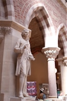 A detailed marble statue of a historical figure stands prominently on a pedestal, set within an archway of an ornate building. The background features decorative brickwork and stone columns. A person wearing a colorful outfit and hat is visible in the background, observing items on display.
