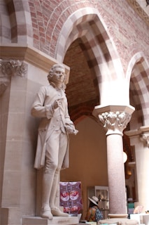 A detailed marble statue of a historical figure stands prominently on a pedestal, set within an archway of an ornate building. The background features decorative brickwork and stone columns. A person wearing a colorful outfit and hat is visible in the background, observing items on display.