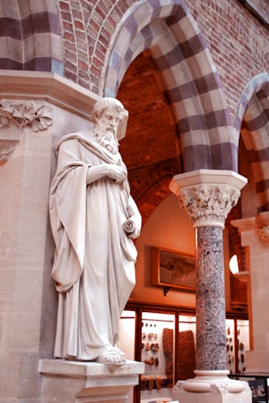 A classical marble statue stands in front of an ornate architectural setting with brick arches and a decorated column. The statue depicts a robed figure with a beard, holding a scroll. The setting includes display cases containing artifacts and a framed picture on the wall, suggesting a museum or historical context.