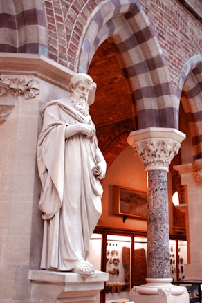 A classical marble statue stands in front of an ornate architectural setting with brick arches and a decorated column. The statue depicts a robed figure with a beard, holding a scroll. The setting includes display cases containing artifacts and a framed picture on the wall, suggesting a museum or historical context.