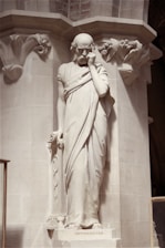 A solemn, bone-white marble statue of a human figure with deep crimson veins subtly glowing beneath the surface, set against a charcoal background.