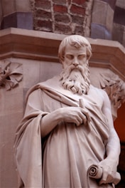 A classical statue of a bearded man draped in a toga, holding a scroll in one hand, carved from stone. The backdrop consists of a combination of stone and brickwork, suggesting an architectural setting.