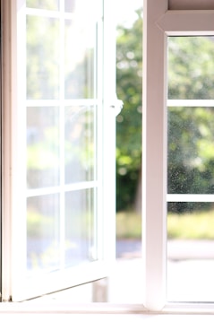An open window with a white frame allowing sunlight to flood through, revealing a lush, green, blurred background outside. The scene conveys a sense of warmth and freshness with natural light bouncing off the window frame, creating a bright and inviting atmosphere.