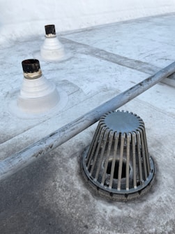 A rooftop surface with a metal drain cover and pipes protruding from the surface. The drain cover has vertical slats surrounding it. The surface appears to be slightly worn with some darker patches and dirt marks.