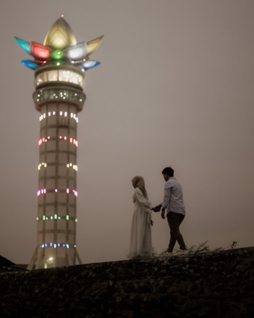 A couple stands hand in hand on a stone platform against the backdrop of a tall, illuminated tower with colorful lights at the top. The scene is set at dusk, creating a moody and romantic atmosphere.